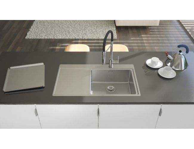 Sodobna kuhinja z belimi omaricami in temnim pultom, v katerega je vgrajeno Alveus Pure inox pomivalno korito z visoko armaturo; na pultu so zložena mapa, skodelici s krožničkoma in kovinski čajnik, v ozadju pa odprt bivalni prostor.